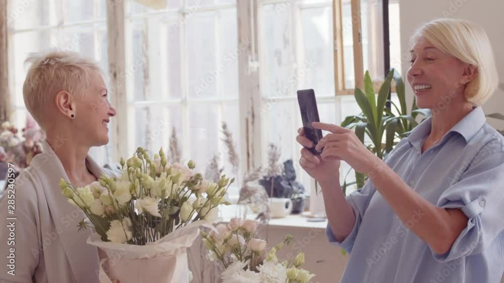Medium shot of blonde middle-aged woman standing with smartphone in her hands and making photos of her female friend standing with flowers in her hands Medium shot of blonde middle-aged woman standing with smartphone in her hands and making photos of her female friend standing with flowers in her hands