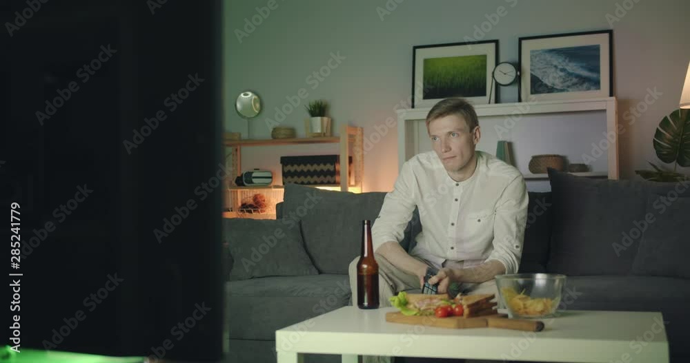 Bored guy is clicking remote control looking for interesting program on TV at night sitting on couch alone with disappointed face. Youth and television concept. Bored guy is clicking remote control looking for interesting program on TV at night sitting on couch alone with disappointed face. Youth and television concept.