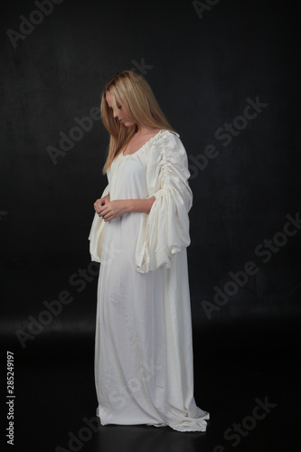 full length portrait of blonde girl wearing long white flowing robe. standing pose against a black studio background. photo