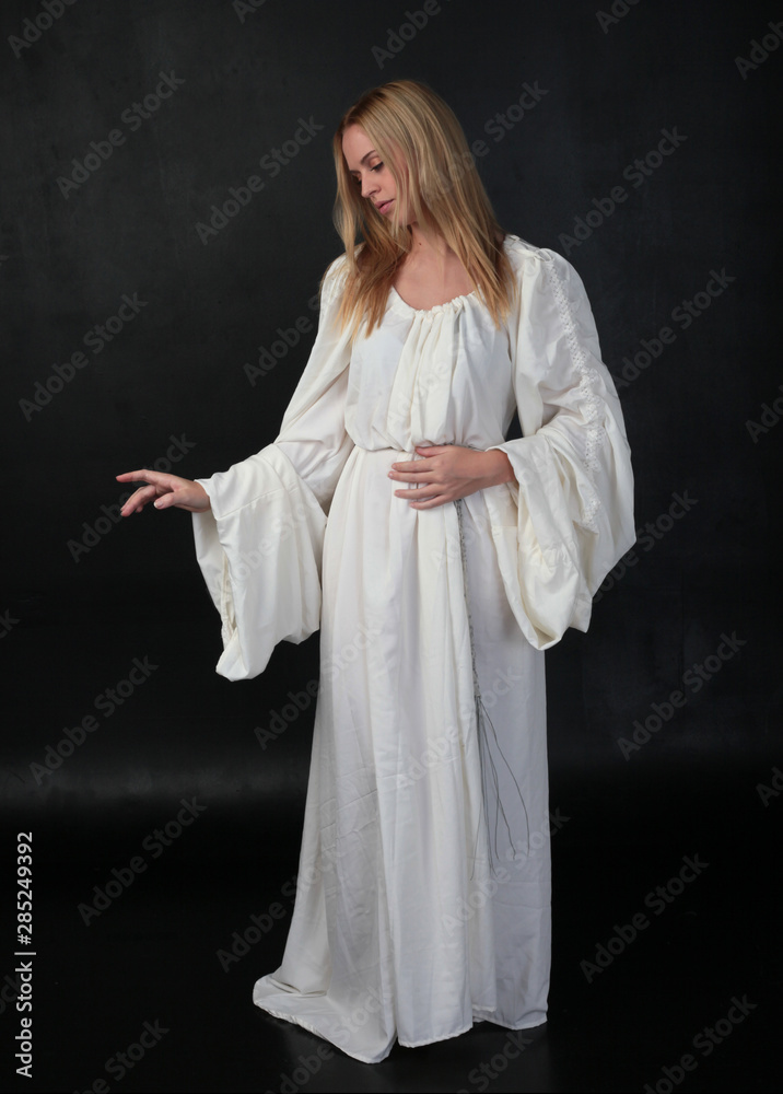 full length portrait of blonde girl wearing long white flowing robe. standing pose against a black studio background. full length portrait of blonde girl wearing long white flowing robe. standing pose against a black studio background.