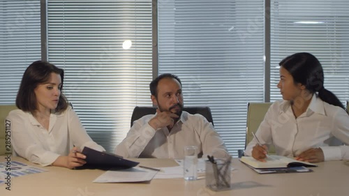 Chest-up shot of tired Caucasian male executive in unbuttoned white shirt sitting in office late at night together with two female associates, who are proposing ideas and trying to persuade him