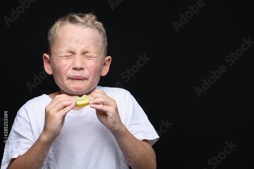 Little, blond boy is eating a piece of a lemon in front of black background and making a facial expression.