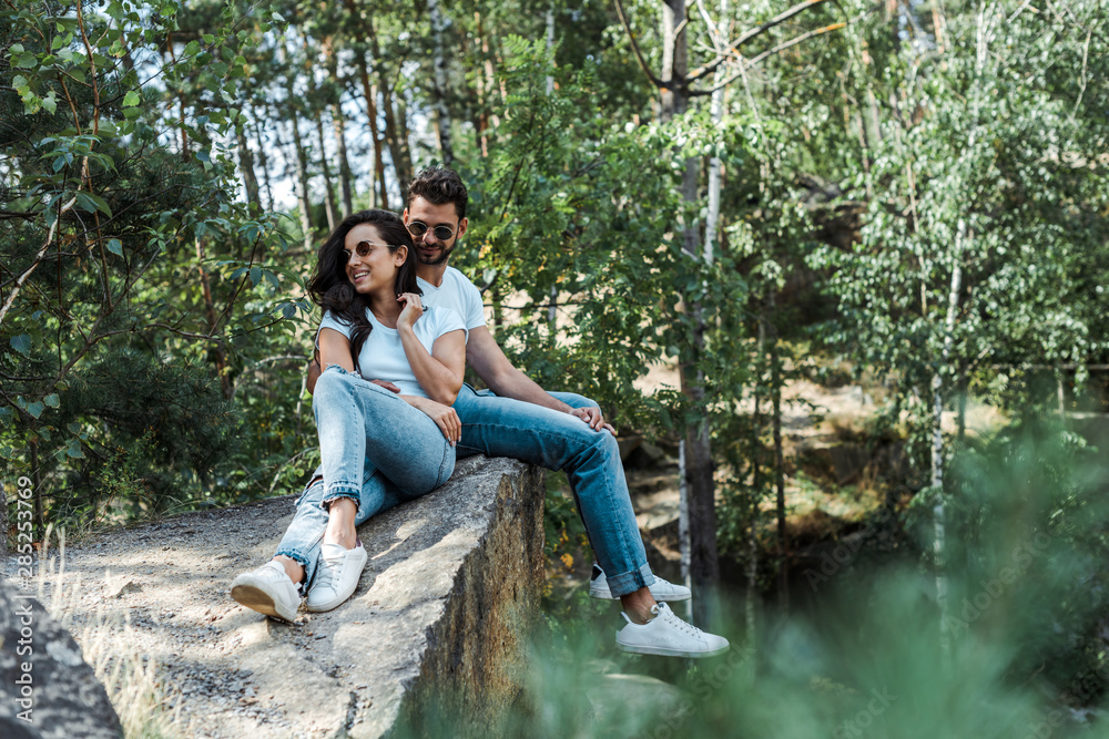 selective focus of handsome man and cheerful woman in sunglasses sitting near trees with leaves selective focus of handsome man and cheerful woman in sunglasses sitting near trees with leaves