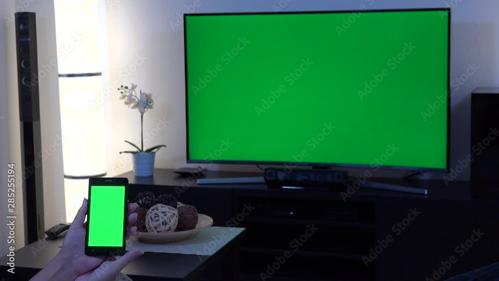 A woman holds out a smartphone with green screen in front of camera in a living room with a TV with green screen