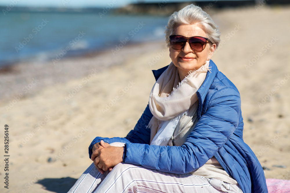 people and leisure concept - happy senior woman in sunglasses and jacket on beach in estonia