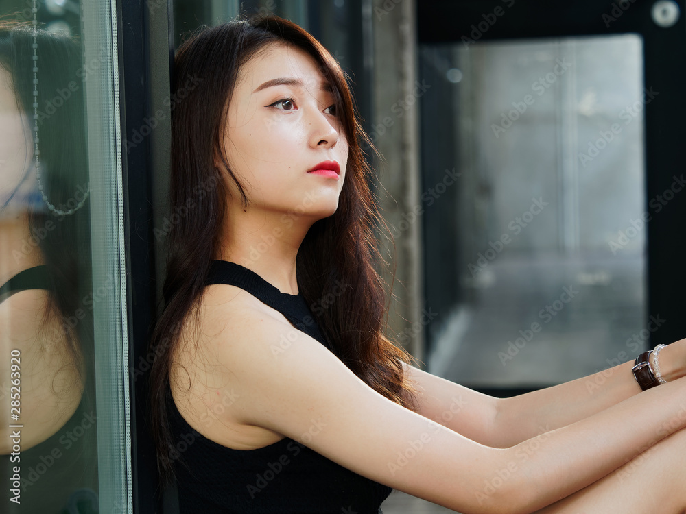 Graceful young woman with long black hair sitting on ground and leaning on the glass wall in suuny day. Outdoor portrait of cool Chinese girl in trendy summer sexy black dress, summer concept. Graceful young woman with long black hair sitting on ground and leaning on the glass wall in suuny day. Outdoor portrait of cool Chinese girl in trendy summer sexy black dress, summer concept.