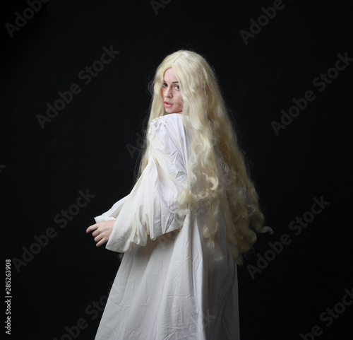 Ghostly portrait of a woman with long blonde hair wearing a white robe. posing against a black studio background. 