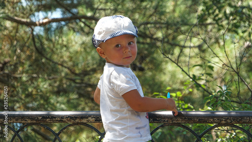 Cute half-standing child holding on to a fence. Trees on background.