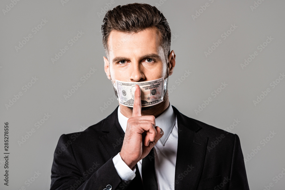 businessman in black suit with dollar banknote on mouth showing hush gesture isolated on grey businessman in black suit with dollar banknote on mouth showing hush gesture isolated on grey