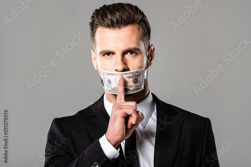 businessman in black suit with dollar banknote on mouth showing hush gesture isolated on grey