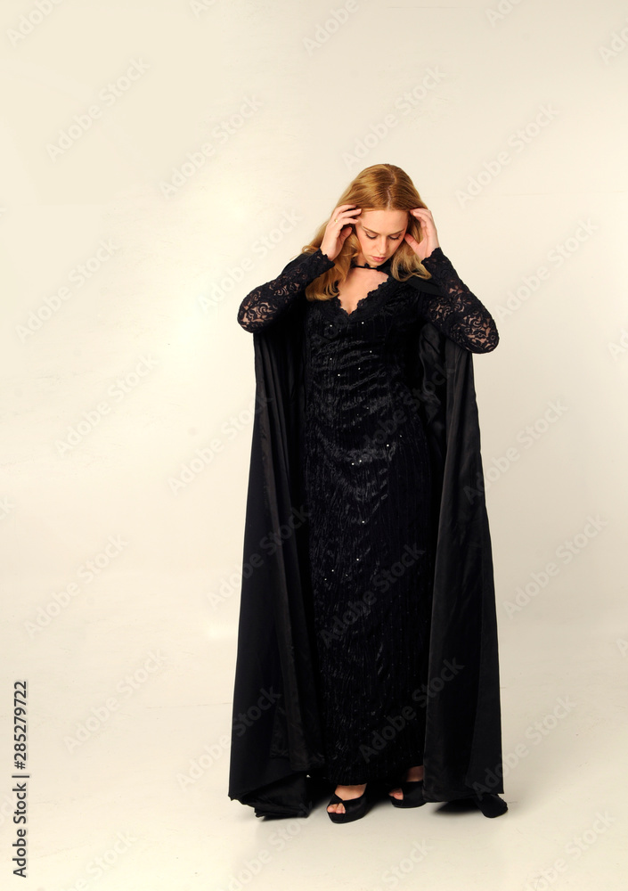 full length portrait of blonde woman wearing long black flowing clock and lace gown, standing pose against a creamy studio background.