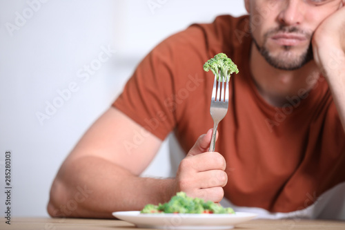 Unhappy man with broccoli on fork at table, closeup photo