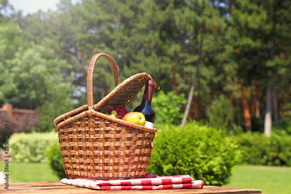Picnic basket with fruits, bottle of wine and checkered blanket on wooden table in garden. Space for text Picnic basket with fruits, bottle of wine and checkered blanket on wooden table in garden. Space for text