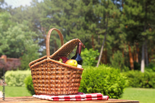 Picnic basket with fruits, bottle of wine and checkered blanket on wooden table in garden. Space for text