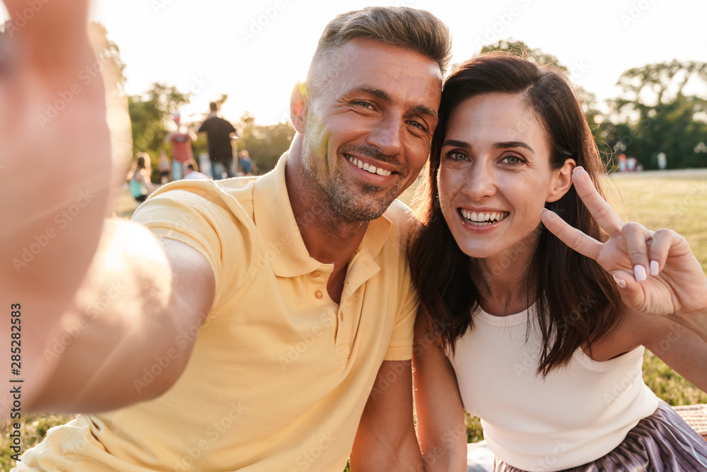 Cheery positive cute adult loving couple outdoors in a beautiful green nature park take a selfie by camera showing peace gesture.