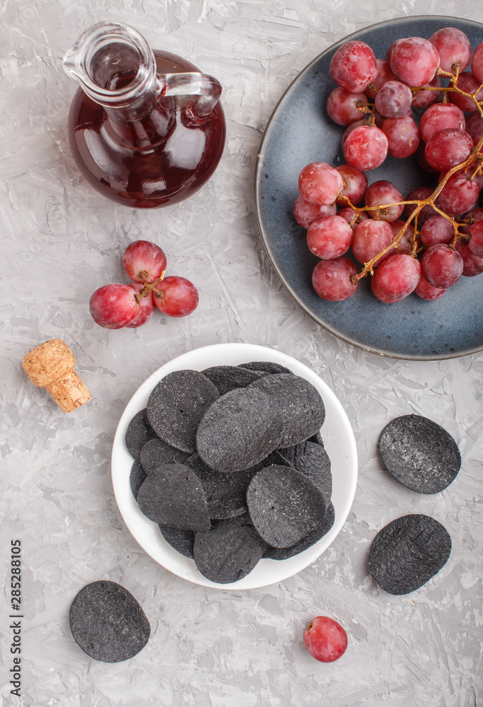 Black potato chips with charcoal, balsamic vinegar in glass, red grapes on a blue ceramic plate on a gray concrete background. Top view. Black potato chips with charcoal, balsamic vinegar in glass, red grapes on a blue ceramic plate on a gray concrete background. Top view.