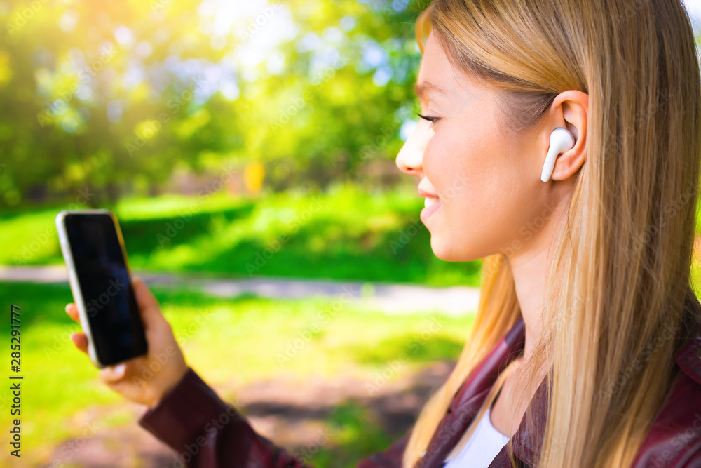 Young beautiful smiling adult Caucasian girl walking outdoor in the park having chat communication with wireless headphones mobile reading talking listening using with her hand on sunset at summer