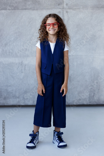 Frontal portrait of a pretty smiling little girl in blue uniform, in red eyeglass, walking with attitude, looking at camera, over concrete background, back to school concept. photo