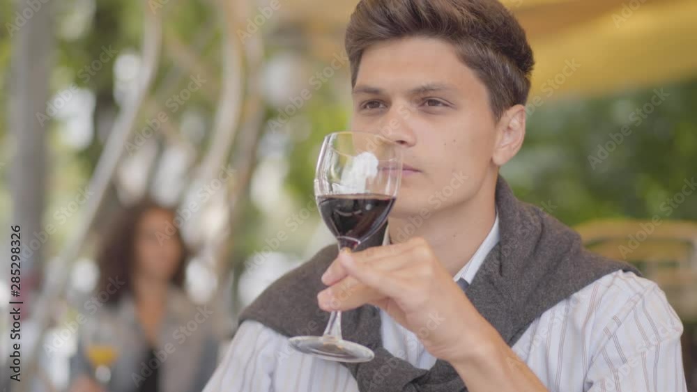 Handsome young man drinking red wine at street cafe. The guy enjoying alcohol relaxing outdoors. The customer sitting at the table tasting beverage. Leisure, day off. Handsome young man drinking red wine at street cafe. The guy enjoying alcohol relaxing outdoors. The customer sitting at the table tasting beverage. Leisure, day off.