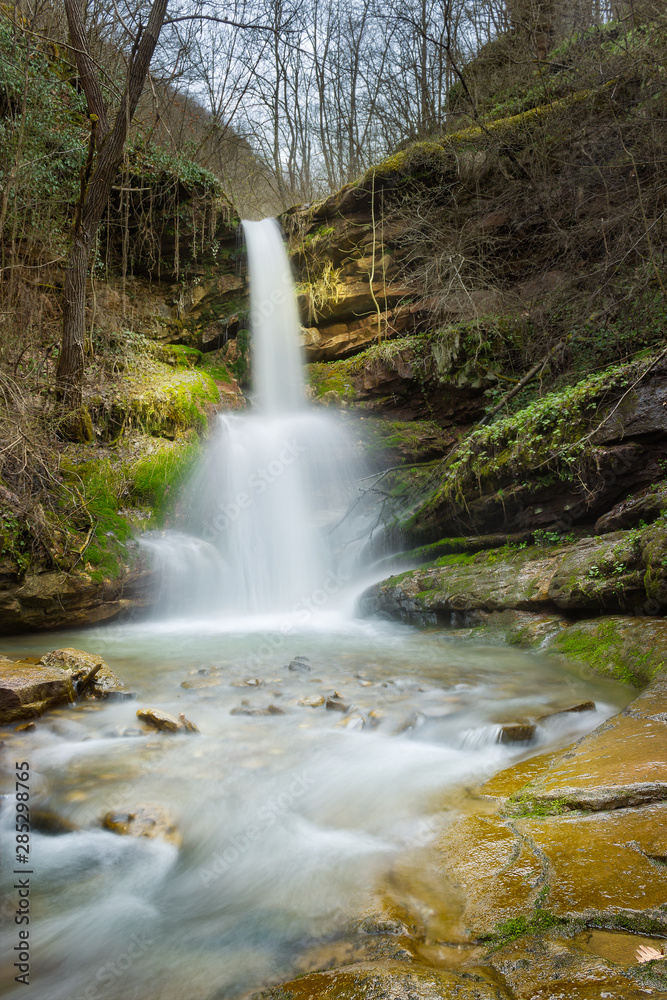 Fototapeta premium Sunlit, colorful forest waterfall streaming down the red rocks in it's amphitheater during spring