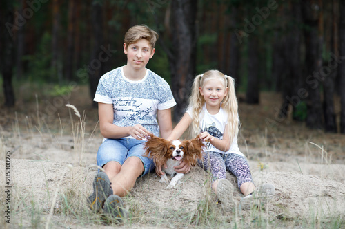Girl with blond hair and a boy are sitting in the forest with their spaniel dog © Dmitry