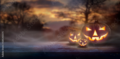Mist rolling in over the spooky glow of Jack O' Lanterns on the right hand side of a wooden bench in a forest at dusk.