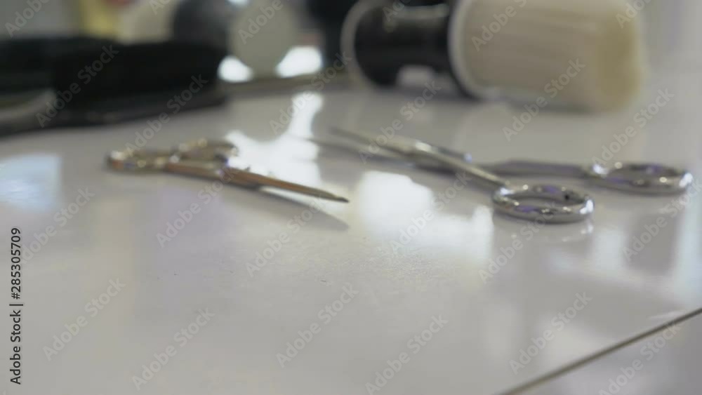 Barber master gently takes a sharp straight razor from a white dresser. Hairstylist tools in the barbershop. Haircutting scissors, hairbrush, straight blade near the mirror on hairdresser stand. Barber master gently takes a sharp straight razor from a white dresser. Hairstylist tools in the barbershop. Haircutting scissors, hairbrush, straight blade near the mirror on hairdresser stand.