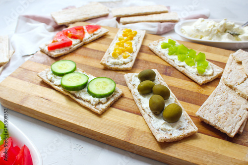 Homemade Crispbread toast with Cottage Cheese and green olives,slices of cabbage,tomatoes,corn,green pepper on cutting board on white concrete background.Healthy food concept,Top view.Flat Lay