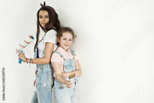 The girls paint the walls. Friendly collaboration between the two sisters. Girlfriends with roller and brush in hands on white wall background. Teens in blue overalls make repairs together.
