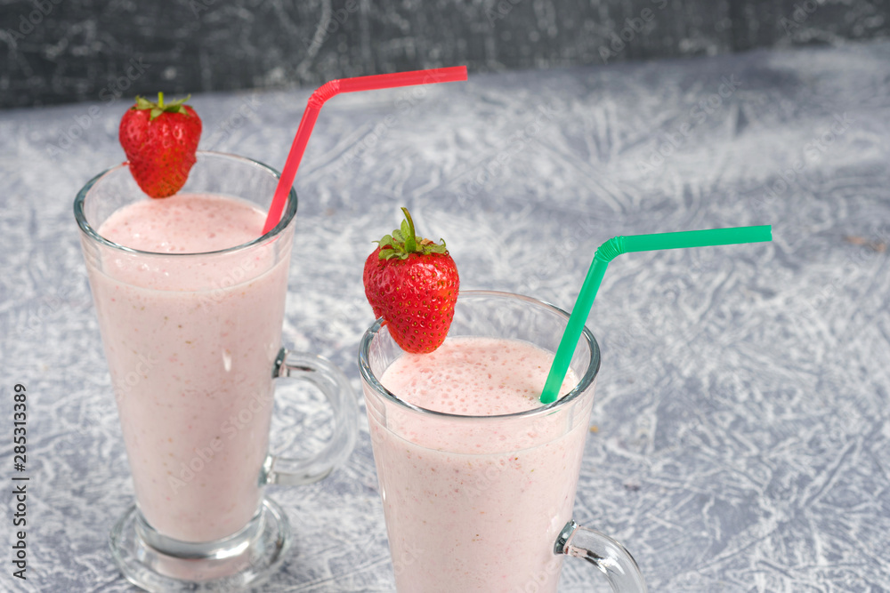 Two glasses with strawberry milkshake with pieces of berries on the edge of the glass on gray background. Two glasses with strawberry milkshake with pieces of berries on the edge of the glass on gray background.