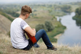 Young boy sits on the hill above the river and plays the guitar