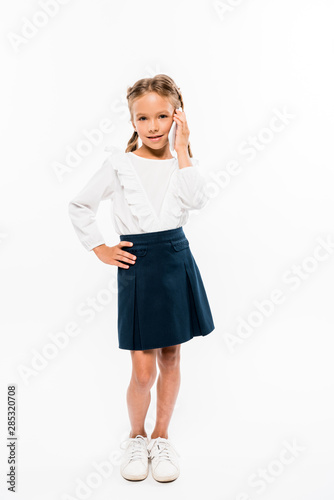 happy kid standing with hand on hip and talking on smartphone isolated on white
