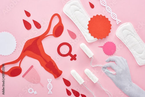 cropped view of white hand holding tampon on pink background with sanitary napkins, paper cut female reproductive internal organs and blood drops photo