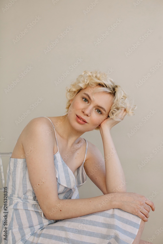 Amazing blond curly girl posing in summer striped dress sitting in studio by the beige background Amazing blond curly girl posing in summer striped dress sitting in studio by the beige background