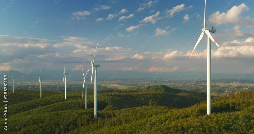 Aerial shot of wind mills rotating by the force of the wind and generating renewable energy in a green ecologic way to the planet with a sun shining.