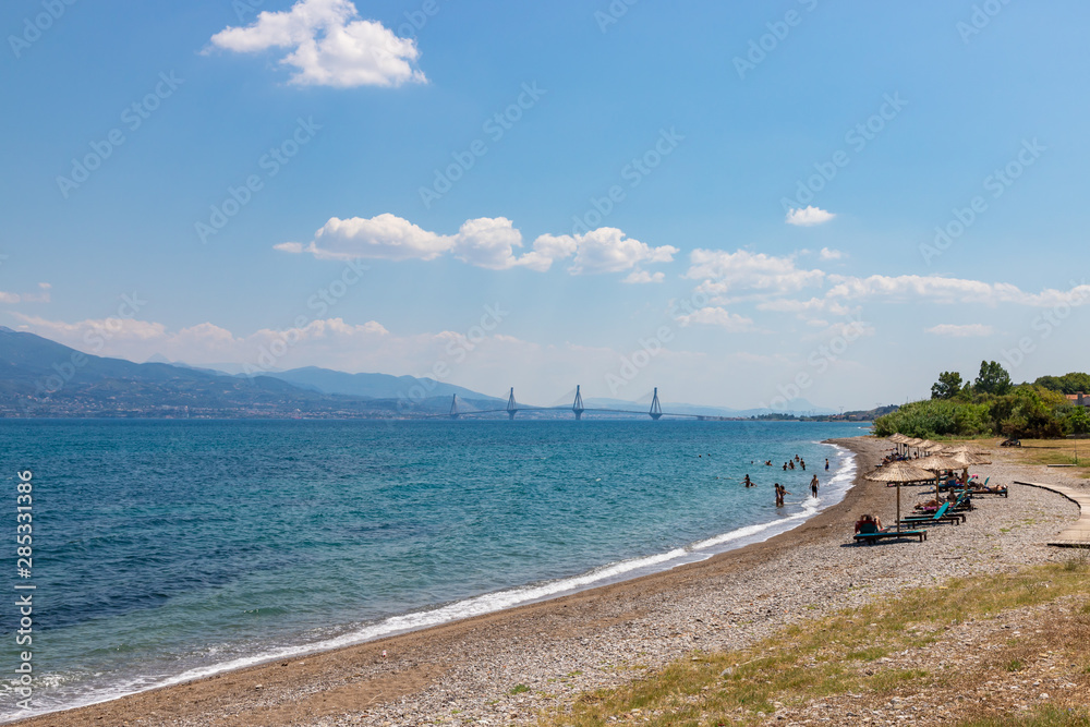 NAFPAKTOS / GREECE - JUNE 27, 2019: People swimming on the beach at Gulf of Corinth  near Nafpaktos town. Rion-Antirion Bridge on the background.