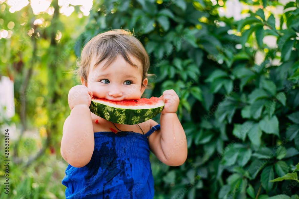 Charming little girl eating watermelon in the yard on the background of plants Charming little girl eating watermelon in the yard on the background of plants