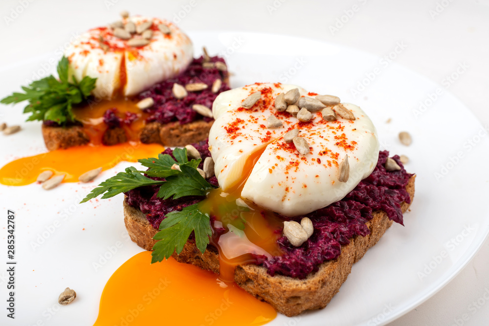 Poached eggs with homemade beetroot hummus, seeds and parsley on toast bread on a white plate on the white background. Healthy breakfast or snack Poached eggs with homemade beetroot hummus, seeds and parsley on toast bread on a white plate on the white background. Healthy breakfast or snack
