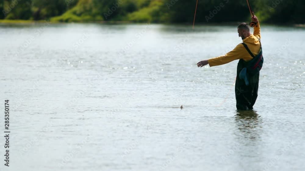 male fisherman in yellow jacket and green special clothes, throws a float, fishing on the river, standing in the water, beautiful nature, small current, summer day sleepy, flying birds, slow motion male fisherman in yellow jacket and green special clothes, throws a float, fishing on the river, standing in the water, beautiful nature, small current, summer day sleepy, flying birds, slow motion