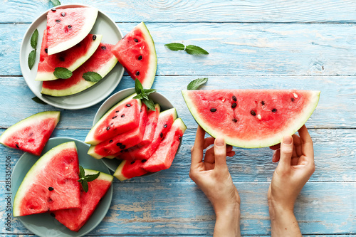 Fresh red watermelon slice in female hands on an blue rustic wood background with copy space. Top view. Summertime concept. Watermelon slice and mint on the background.