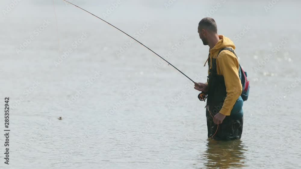 man fisherman in yellow jacket and green special clothes, throws float, caught fish in the river, standing in the water, beautiful nature, small current, summer Sunny day, birds fly, wide angle man fisherman in yellow jacket and green special clothes, throws float, caught fish in the river, standing in the water, beautiful nature, small current, summer Sunny day, birds fly, wide angle