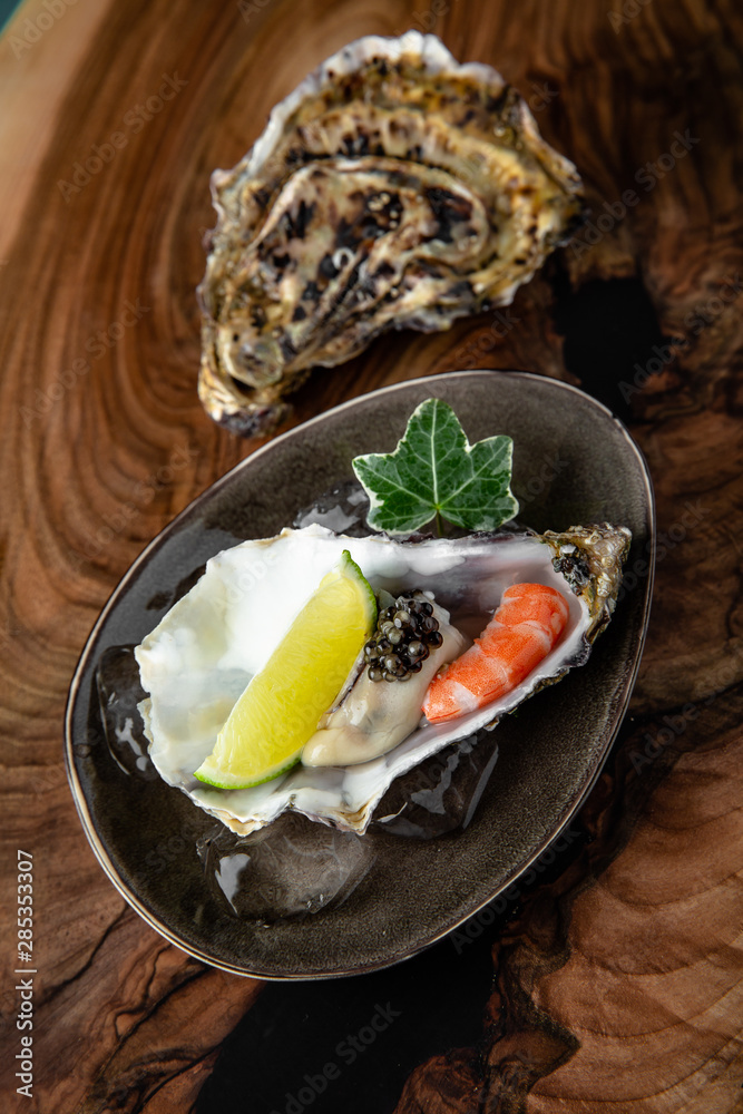 Fresh oysters with black sturgeon caviar and shrimp in ice, with lime in a luxurious serve in a ceramic plate on a textured wooden table.