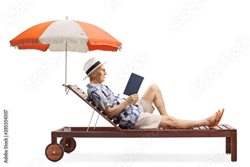 Canvas Print Senior male tourist reading a book on a sunbed under an umbrella