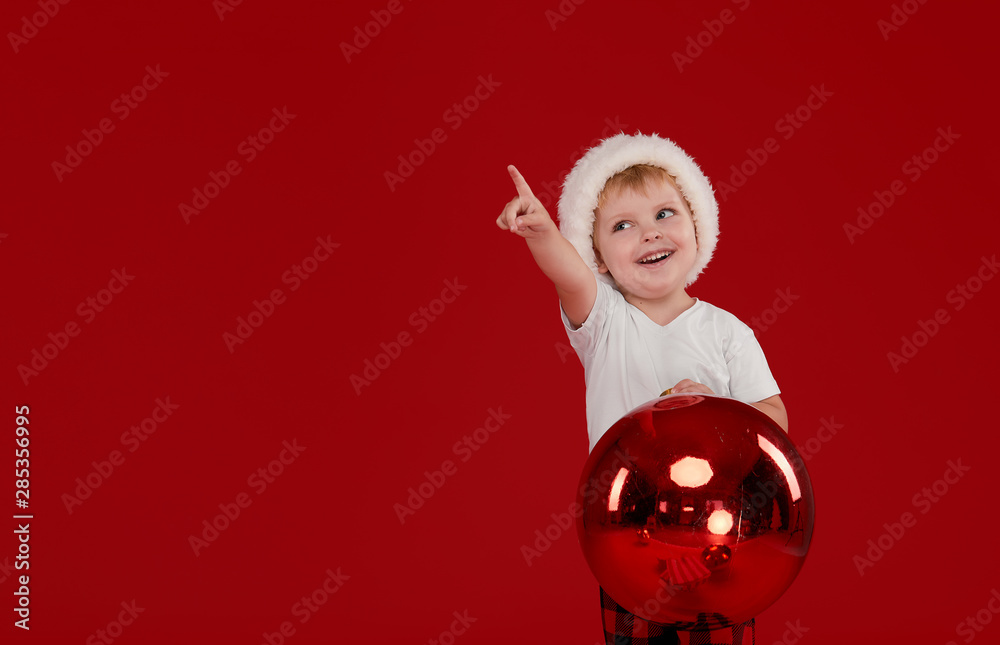 Little kid boy in santa claus hat holds big christmas ball on red isolated background. Point finger left, blank space for text.