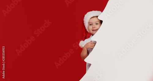 Happy little girl child in Santa hat peeking from behind blank sign banner in the shape of Christmas trees. Isolated on red background. Space for Your Text. Sale, holidays, christmas, new year concept