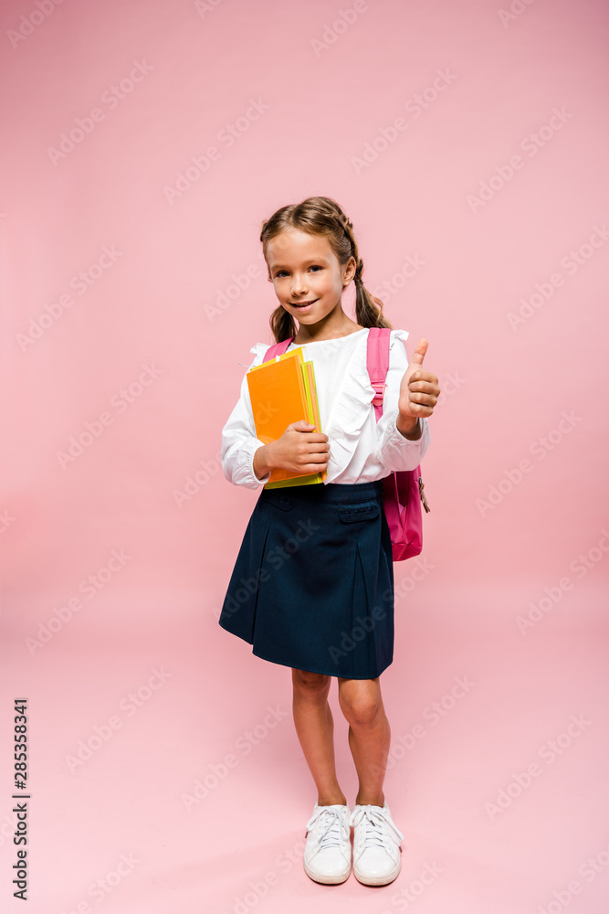happy kid holding books while standing with backpack and showing thumb up on pink happy kid holding books while standing with backpack and showing thumb up on pink