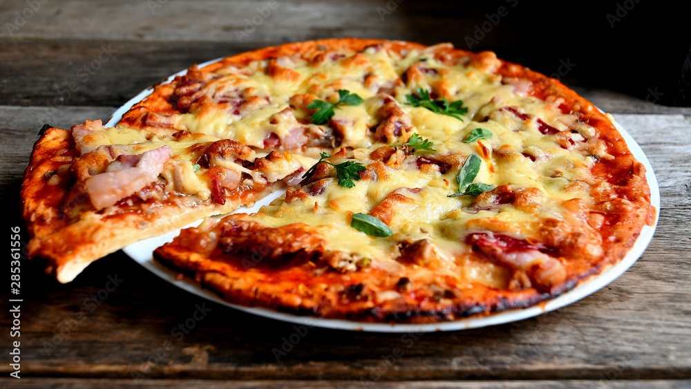Homemade pizza with cheese, tomatoes, bacon and herbs in a white ceramic plate on a wooden table.
