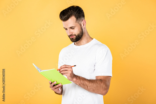 handsome man in white t-shirt writing in notebook isolated on yellow
