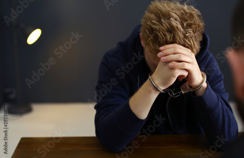 Man detained in handcuffs at desk indoors, space for text. Criminal law photo