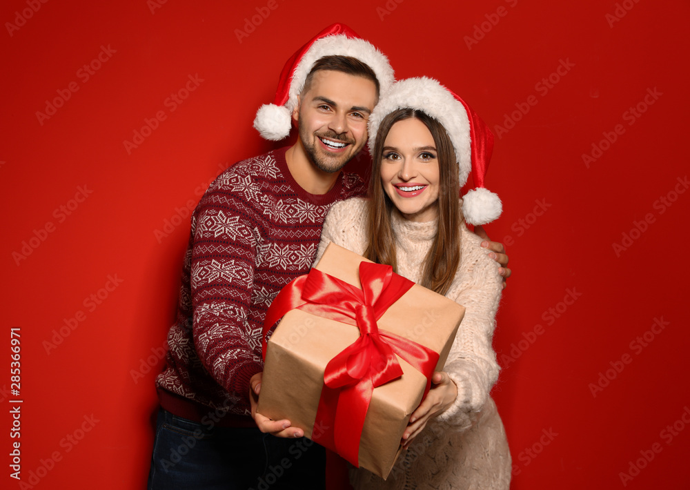 Couple in Christmas sweaters and Santa hats with gift box on red background Couple in Christmas sweaters and Santa hats with gift box on red background